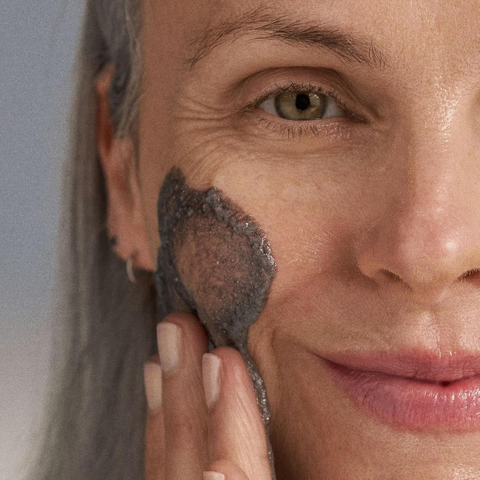 Woman applying a dark gray facial mask to her cheek against a neutral background
