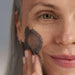 Woman applying a dark gray facial mask to her cheek against a neutral background