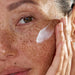 Close-up of a person applying cream to their face with visible freckles.